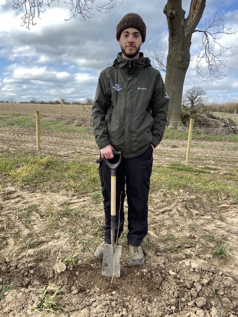 George, Apprentice Forest Operative, stands in a field looking at the camera. He is dressed for cold weather and holding a spade.