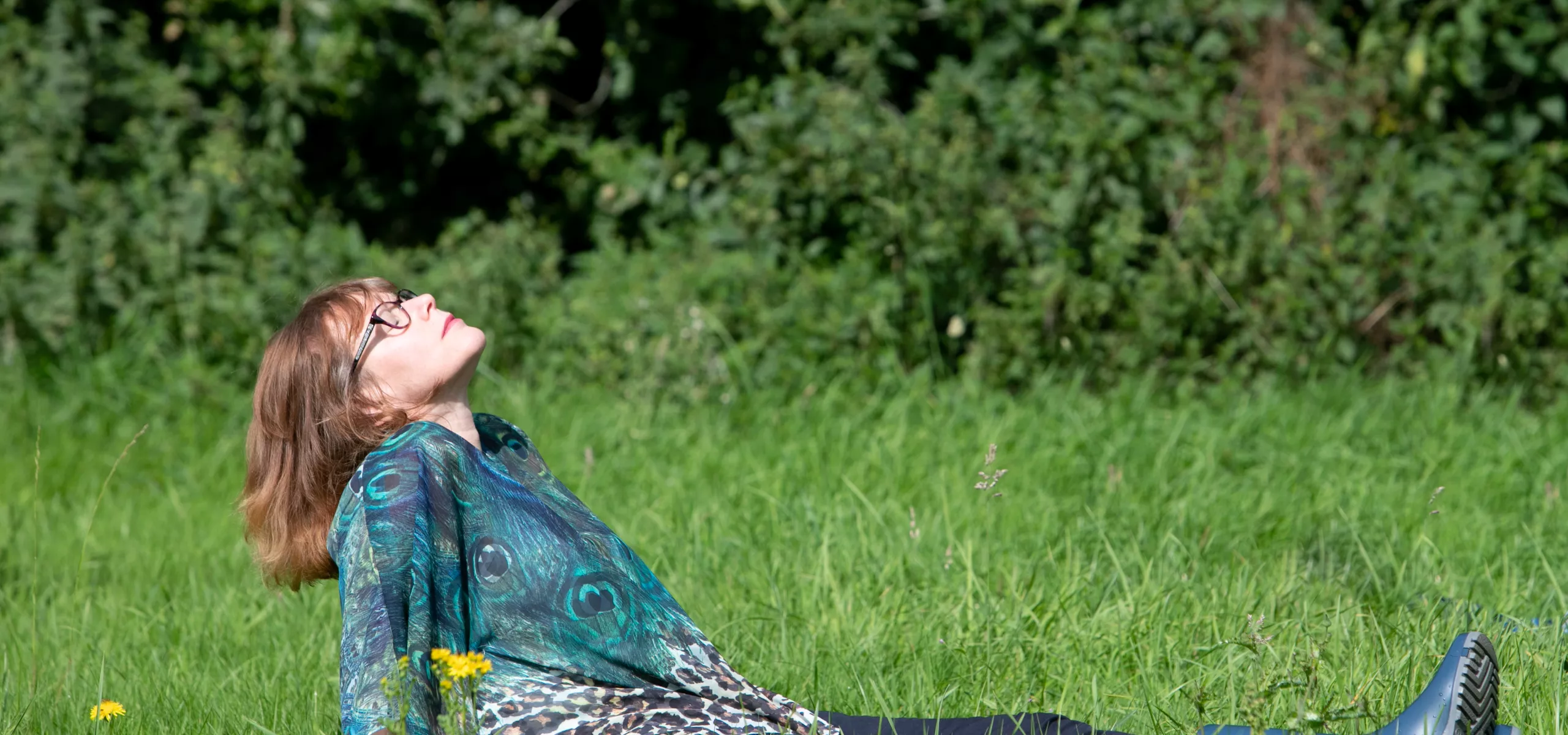A person sat in the grass, leaning back in a relax pose with their head tiled towards the sky and their eyes closed.
